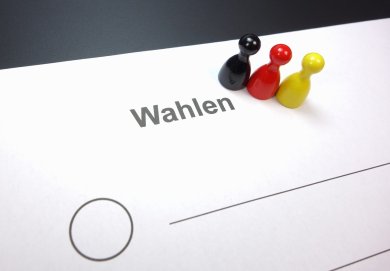 Drei Spielfiguren - schwarz, rot, gelb - stehen neben dem Wort "Wahlen" - Symbolbild