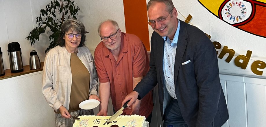 Eine Frau und zwei Männer hinter einem Tisch, auf dem eine Torte steht mit der Aufschrift 15 Jahre Zeitraum