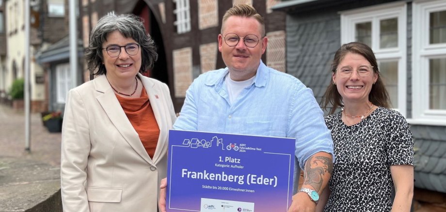 Drei Personen mit einer Gewinnerplakette vor dem historischen Rathaus
