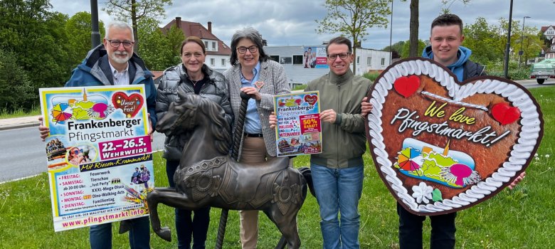 Menschengruppe um ein bronzenes Karussellpferd mit Plakaten und Ankündigungen für den Pfingstmarkt