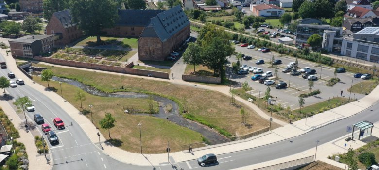 Luftbild mit renaturiertem Bachlauf und Parkplatz im Umfeld einer historischen Klosteranlage